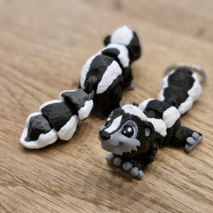 Könnte beinhalten: Zwei Stinktierfiguren, eine liegend und eine aufrecht, werden auf einer Holzoberfläche gezeigt. Die Stinktiere sind schwarz und weiß mit grauen Akzenten auf ihren Gesichtern und Füßen. Das liegende Stinktier hat einen Schlüsselanhänger.