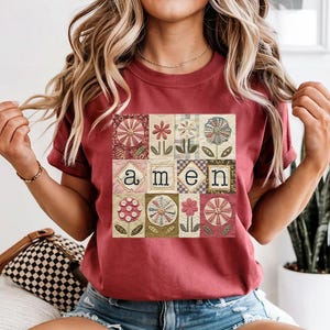 May include: A maroon t-shirt featuring a patchwork design with floral and geometric patterns. The word "amen" is centered within the design. The shirt has short sleeves and a crew neck.
