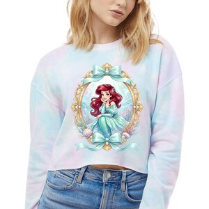 Puede incluir: Sudadera corta te&ntilde;ida con un dise&ntilde;o de sirena de pelo rojo y vestido turquesa enmarcada en oro con lazos turquesa. Conchas marinas y plantas acu&aacute;ticas rodean el marco. La sudadera es de color pastel.