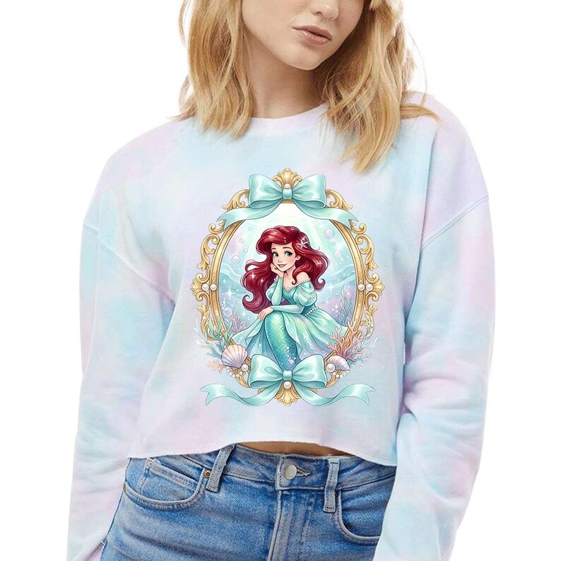 Puede incluir: Sudadera corta te&ntilde;ida con un dise&ntilde;o de sirena de pelo rojo y vestido turquesa enmarcada en oro con lazos turquesa. Conchas marinas y plantas acu&aacute;ticas rodean el marco. La sudadera es de color pastel.