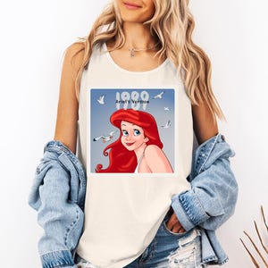 May include: Cream-colored tank top featuring a square graphic of Ariel from The Little Mermaid. The graphic includes the text "1989 Ariel's Version" with Ariel's red hair and a blue background with seagulls. The tank top is sleeveless.