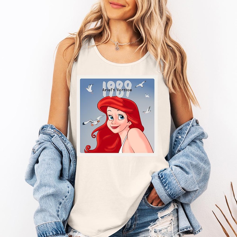May include: Cream-colored tank top featuring a square graphic of Ariel from The Little Mermaid. The graphic includes the text "1989 Ariel's Version" with Ariel's red hair and a blue background with seagulls. The tank top is sleeveless.