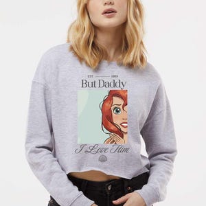 May include: A gray cropped sweatshirt with the text "But Daddy" above a cartoon image of a red-haired character and the words "I Love Him" below. The sweatshirt has a raw hem and long sleeves.
