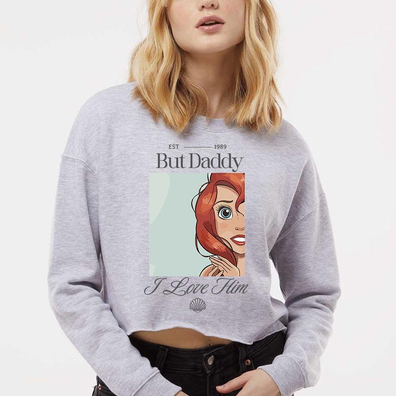 May include: A gray cropped sweatshirt with the text "But Daddy" above a cartoon image of a red-haired character and the words "I Love Him" below. The sweatshirt has a raw hem and long sleeves.