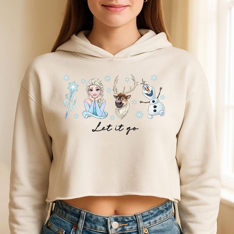 May include: A cream-coloured cropped hoodie with a graphic of characters from the film Frozen, including Elsa, Olaf, and Sven, with the phrase "Let it go" in black script. The design includes snowflakes and a blue magic wand.