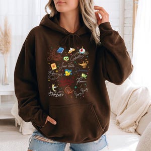May include: A dark brown hooded sweatshirt featuring a front graphic print of various colorful icons and cursive names including Belle, Cinderella, Snow White, Jasmine, Fiona, Rapunzel, and Tinker Bell, along with small star accents and a front pocket.