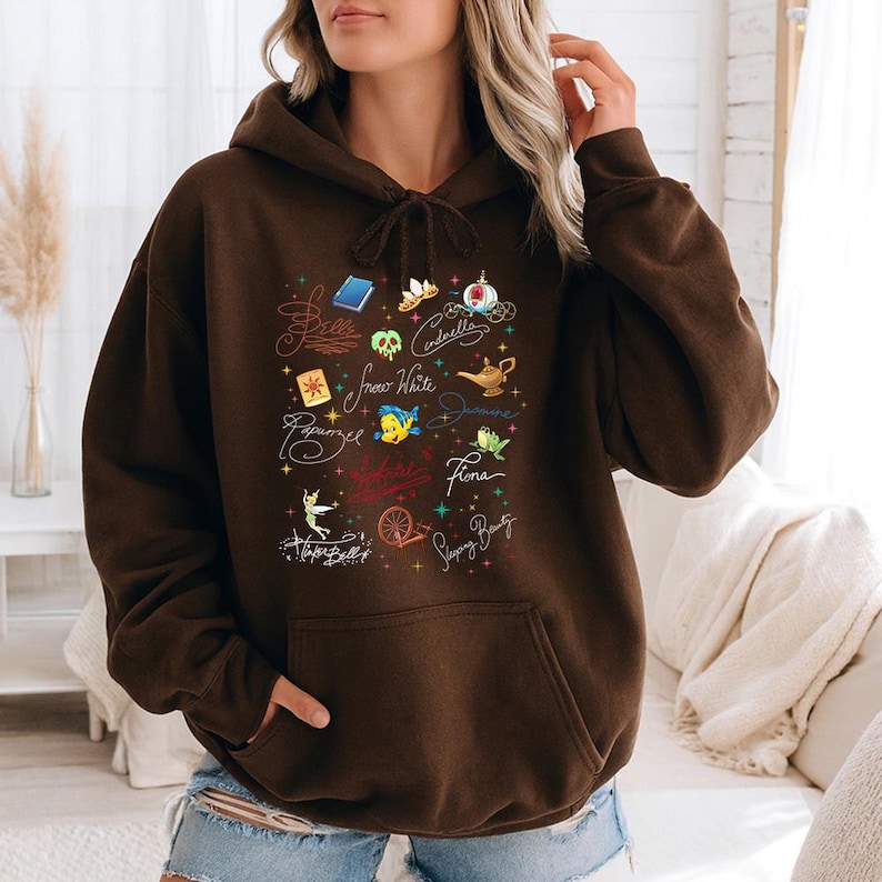 May include: A dark brown hooded sweatshirt featuring a front graphic print of various colorful icons and cursive names including Belle, Cinderella, Snow White, Jasmine, Fiona, Rapunzel, and Tinker Bell, along with small star accents and a front pocket.