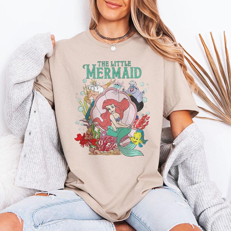 May include: A beige short-sleeve t-shirt featuring a graphic print of The Little Mermaid characters including Ariel, King Triton, Ursula, Sebastian, and Flounder. The design includes colourful underwater elements like coral, seaweed, and bubbles.
