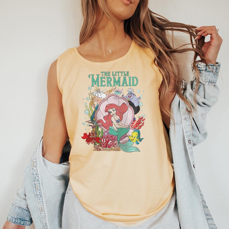 May include: A pale yellow sleeveless tank top featuring a graphic print of The Little Mermaid characters including Ariel, King Triton, Ursula, Sebastian, and Flounder. The design includes the text The Little Mermaid in teal lettering.