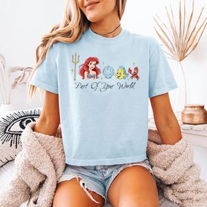 May include: Light blue t-shirt featuring a graphic of Ariel, Flounder, Sebastian, and a seashell, with a golden trident and the text "Part of Your World." The shirt is a comfortable, casual style.