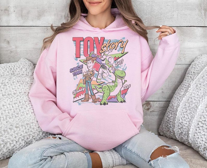 May include: A light pink hooded sweatshirt featuring a graphic print of Toy Story characters Woody, Buzz Lightyear, and Rex. The design includes the text "TOY story", "SINCE 1995", "Woody", "Buzz", and "Rex" with colorful geometric shapes in the background.