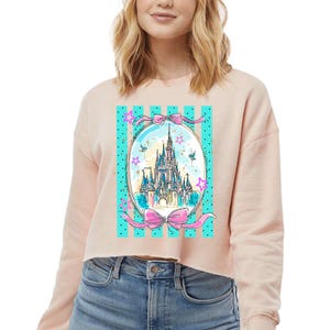 May include: A light pink cropped sweatshirt featuring a graphic of a fairytale castle inside an oval frame with pink bows and turquoise stripes. The castle is detailed with blue, yellow, and pink accents, surrounded by stars and birds.