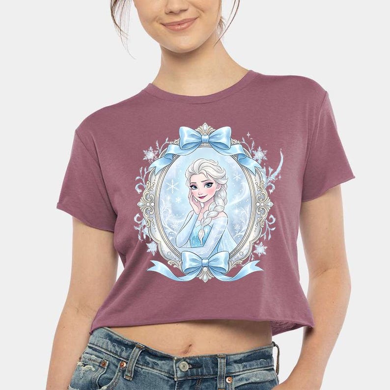 Pode incluir: Um top curto bord&ocirc; com uma ilustra&ccedil;&atilde;o emoldurada de uma princesa, com la&ccedil;os e fitas azuis. A princesa tem cabelo branco e usa um vestido azul. A camisa &eacute; feita de um material macio.