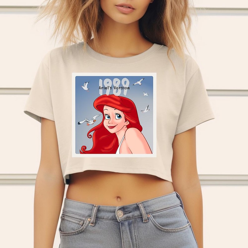 May include: A cream-colored crop top featuring a square graphic of Ariel from The Little Mermaid. The graphic includes the text "1989 Ariel's Version" with Ariel's red hair and blue eyes. The top is paired with blue jeans.