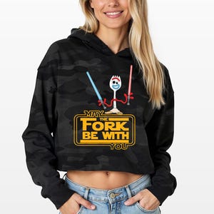 May include: A black camouflage print cropped hoodie featuring a graphic of a white spork character holding a blue and a red lightsaber. Below the character, the yellow text reads MAY THE FORK BE WITH YOU in a stylized rectangular frame.