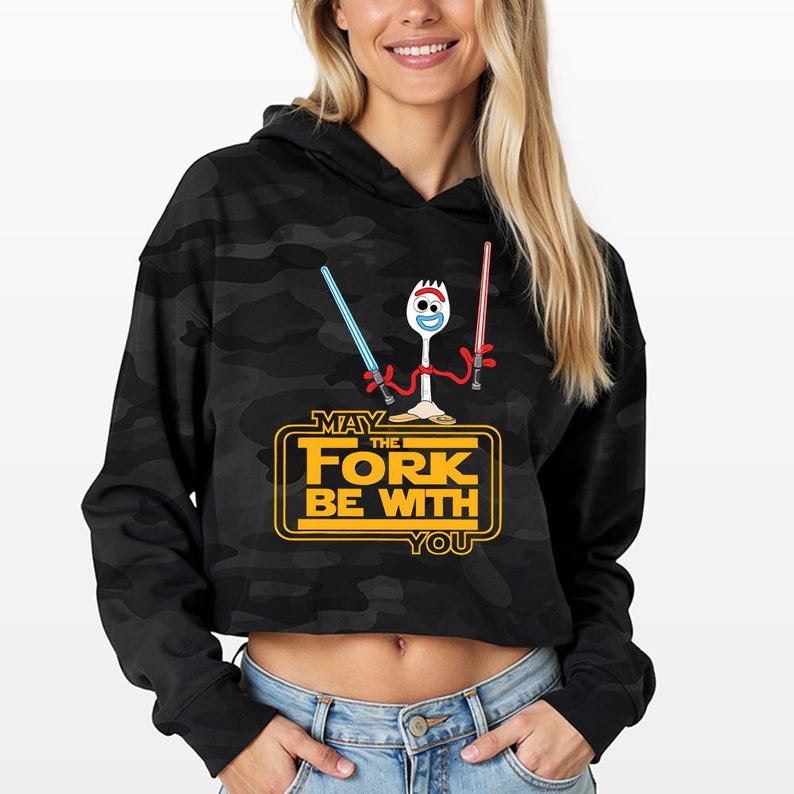 May include: A black camouflage print cropped hoodie featuring a graphic of a white spork character holding a blue and a red lightsaber. Below the character, the yellow text reads MAY THE FORK BE WITH YOU in a stylized rectangular frame.