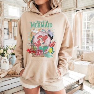 May include: A beige hooded sweatshirt featuring a graphic print of The Little Mermaid characters including Ariel, King Triton, Ursula, Sebastian, and Flounder. The design includes a large pink seashell, red coral, and bubbles with the text The Little Mermaid.