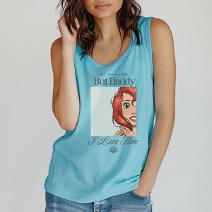 May include: A light blue tank top with a scoop neck. The shirt features a graphic of a cartoon character with red hair and the text "But Daddy" above the image and "I Love Him" below it.