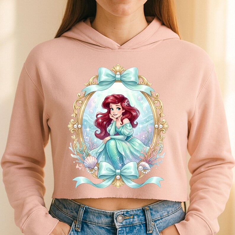 Op de afbeelding: Een lichtroze cropped hoodie met een ingelijste illustratie van een zeemeermin met rood haar en een zeegroene staart. De lijst is goudkleurig met zeegroene strikken. De zeemeermin draagt een zeegroene jurk.