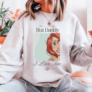 May include: A light gray sweatshirt featuring the text "But Daddy" above an illustration of a cartoon character with red hair and the text "I Love Him". The sweatshirt has a crew neck.