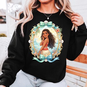 May include: Black sweatshirt featuring a colorful illustration of a woman in a tropical setting. The artwork is framed by an ornate gold oval with bows and floral accents. The woman has long dark hair and is wearing a red and white outfit.