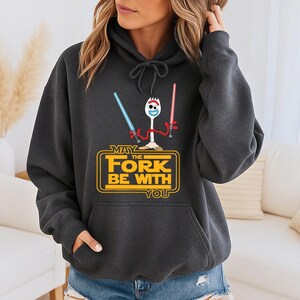 May include: A dark gray hooded sweatshirt featuring a graphic of a white spork character holding a blue and a red light saber. Below the character, yellow text reads MAY THE FORK BE WITH YOU, enclosed in a rectangular border with rounded corners.