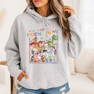 May include: A light gray hoodie featuring a colorful graphic of characters from the movie Toy Story. The graphic includes the text "YOU'VE GOT A FRIEND IN ME" in a playful font, along with images of Woody, Buzz Lightyear, and other characters.