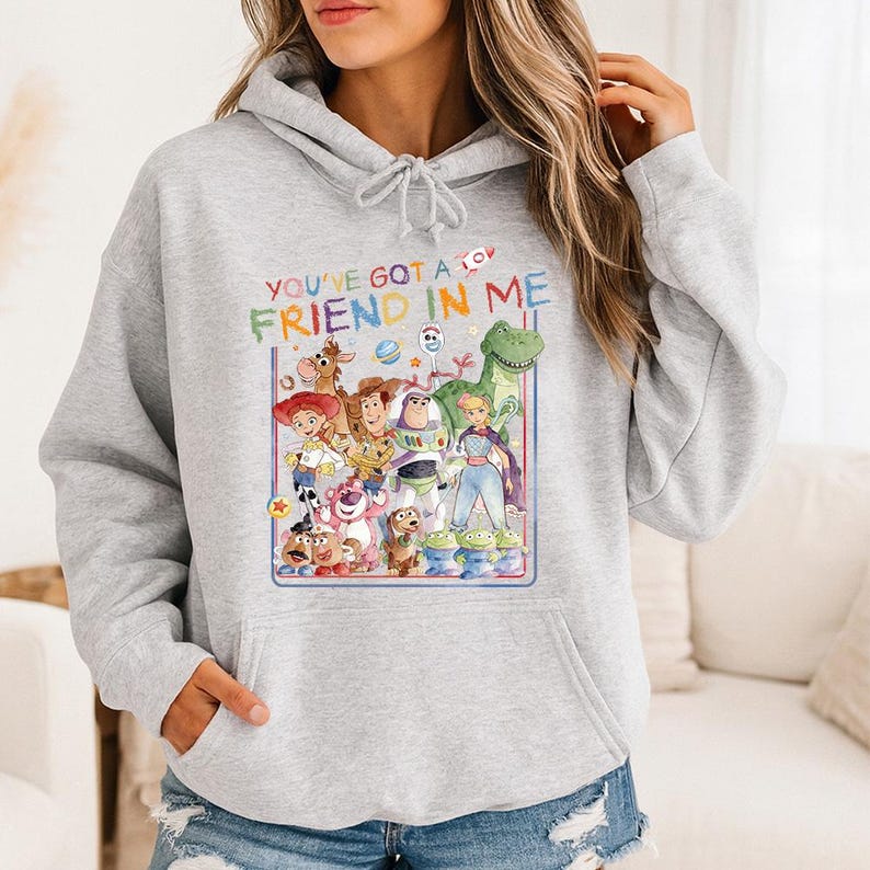 May include: A light gray hoodie featuring a colorful graphic of characters from the movie Toy Story. The graphic includes the text "YOU'VE GOT A FRIEND IN ME" in a playful font, along with images of Woody, Buzz Lightyear, and other characters.