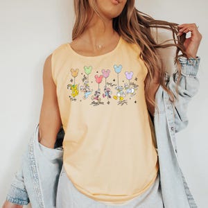 May include: A pale yellow vest top with a vibrant graphic of Disney characters, each holding a Mickey Mouse balloon. Characters include Pluto, Mickey, Minnie, Donald, and Daisy Duck. The text "Mickey Mouse Days" is printed below the characters.