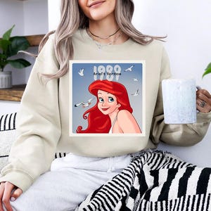 May include: Beige sweatshirt featuring a graphic of Ariel from The Little Mermaid, with the text "1989 Ariel's Version". The image shows Ariel with red hair and seagulls. The sweatshirt is a casual style.