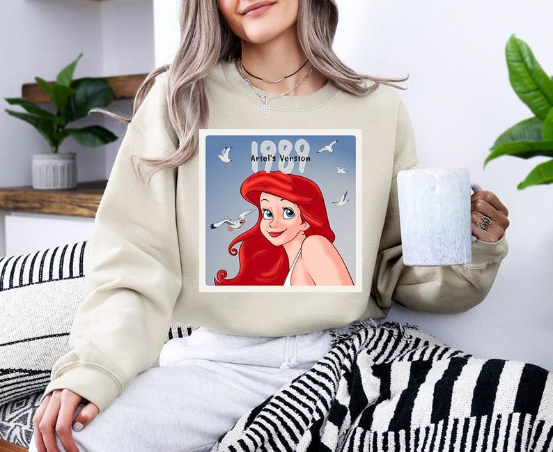 May include: Beige sweatshirt featuring a graphic of Ariel from The Little Mermaid, with the text "1989 Ariel's Version". The image shows Ariel with red hair and seagulls. The sweatshirt is a casual style.