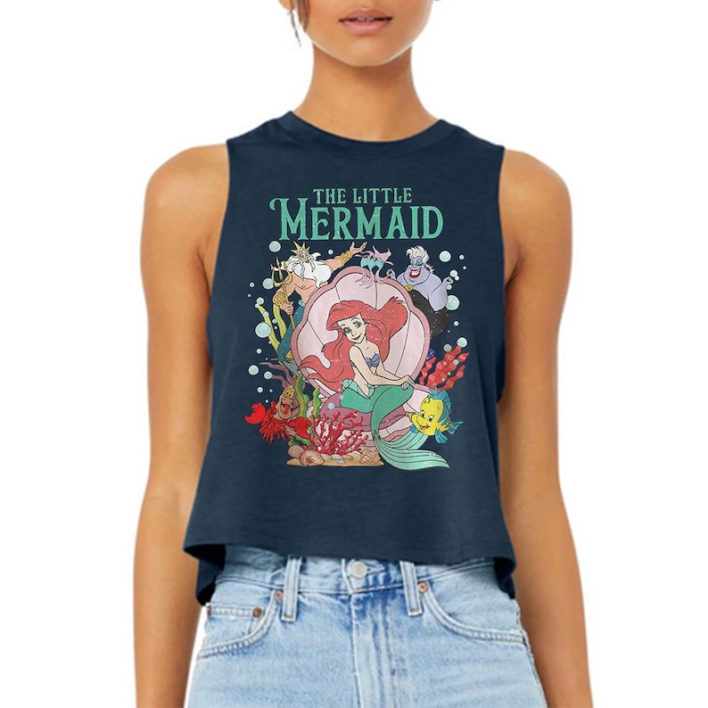 Puede incluir: Un top corto sin mangas de color azul marino oscuro con un estampado gr&aacute;fico de los personajes de La Sirenita, incluyendo a Ariel, el Rey Trit&oacute;n, &Uacute;rsula, Sebasti&aacute;n y Flounder. El texto THE LITTLE MERMAID aparece en verde claro arriba.
