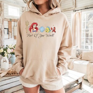 May include: Beige hoodie featuring a graphic of Ariel, Flounder, Sebastian, and a seashell, with a golden trident. The text "Part Of Your World" is below the graphic. The hoodie has a front pocket and a drawstring hood.
