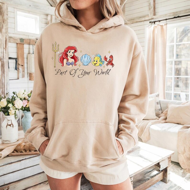 May include: Beige hoodie featuring a graphic of Ariel, Flounder, Sebastian, and a seashell, with a golden trident. The text "Part Of Your World" is below the graphic. The hoodie has a front pocket and a drawstring hood.