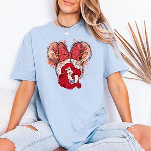 Puede incluir: Camiseta azul claro con un dise&ntilde;o de Ariel de La Sirenita. El dise&ntilde;o incluye un lazo rojo con lunares, detalles florales y el pelo rojo del personaje. La camiseta es de estilo casual.