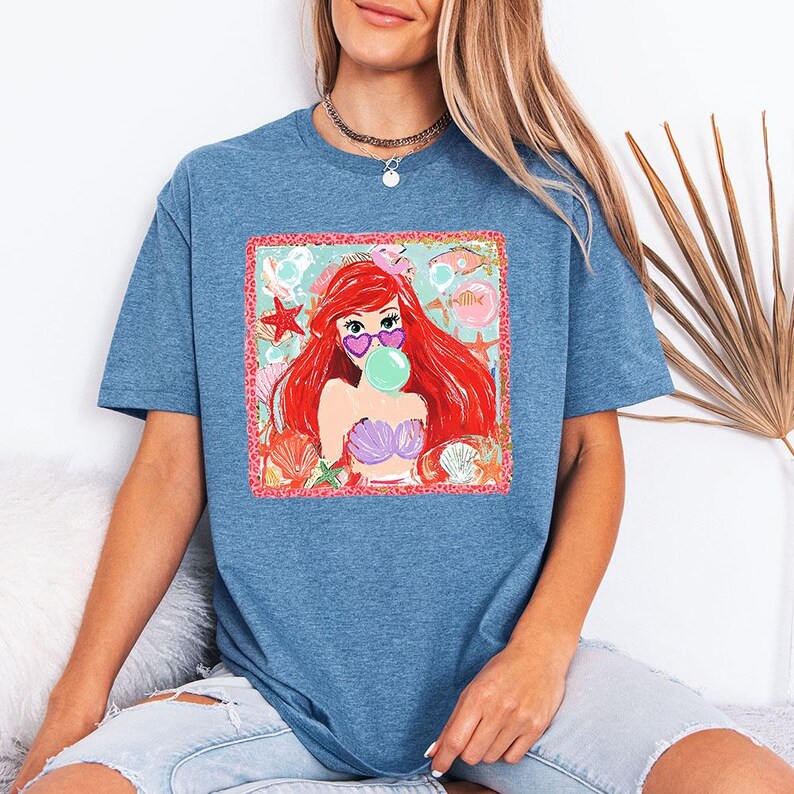 May include: Blue short-sleeved t-shirt featuring a colorful graphic of a mermaid with red hair, heart-shaped sunglasses, and a bubblegum bubble. The graphic includes seashells, starfish, and fish. The shirt is a casual style.