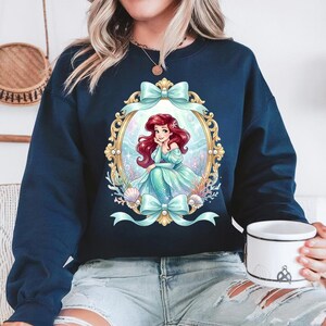 May include: Navy blue sweatshirt featuring a framed illustration of a mermaid with red hair and a teal dress. The frame is gold with teal bows at the top and bottom. The mermaid is in a sea setting with seashells and coral.