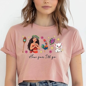 May include: Pale pink crop top with a graphic of a Polynesian woman, a rooster, a pig, and other tropical elements. The text "How far I'll go" is printed below the graphic. The shirt has short sleeves and a crew neck.