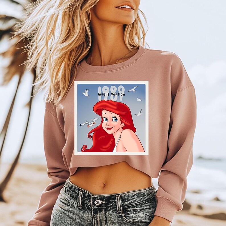 Pode incluir: Um moletom cropped rosa empoeirado com um gr&aacute;fico de Ariel de A Pequena Sereia. O gr&aacute;fico &eacute; uma imagem quadrada com o texto "1989 Ariel's Version" e inclui gaivotas. A modelo est&aacute; usando jeans azul.