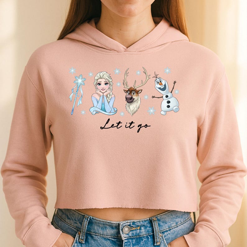 May include: A pale pink cropped hoodie with a graphic of Elsa, Olaf, Sven, and a snowflake wand. The words "Let it go" are written in black script below the characters. The hoodie is worn with blue jeans.