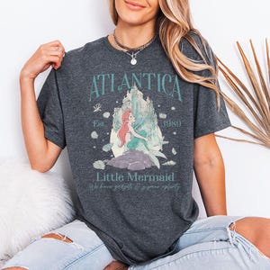 May include: Dark gray t-shirt featuring a graphic of a mermaid with the text "Atlantica" and "Little Mermaid." The design includes a castle, coral, and the year 1989. The shirt also has the phrase "We have gadgets & gizmos aplenty."
