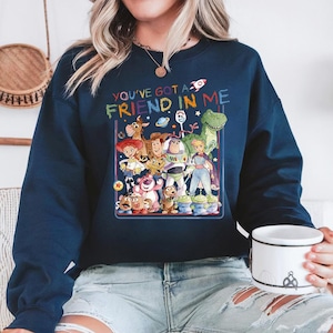 May include: Navy blue sweatshirt featuring a colorful graphic of characters from the animated film "Toy Story." The graphic includes the text "YOU'VE GOT A FRIEND IN ME" in rainbow colors. The sweatshirt is a crew neck style.