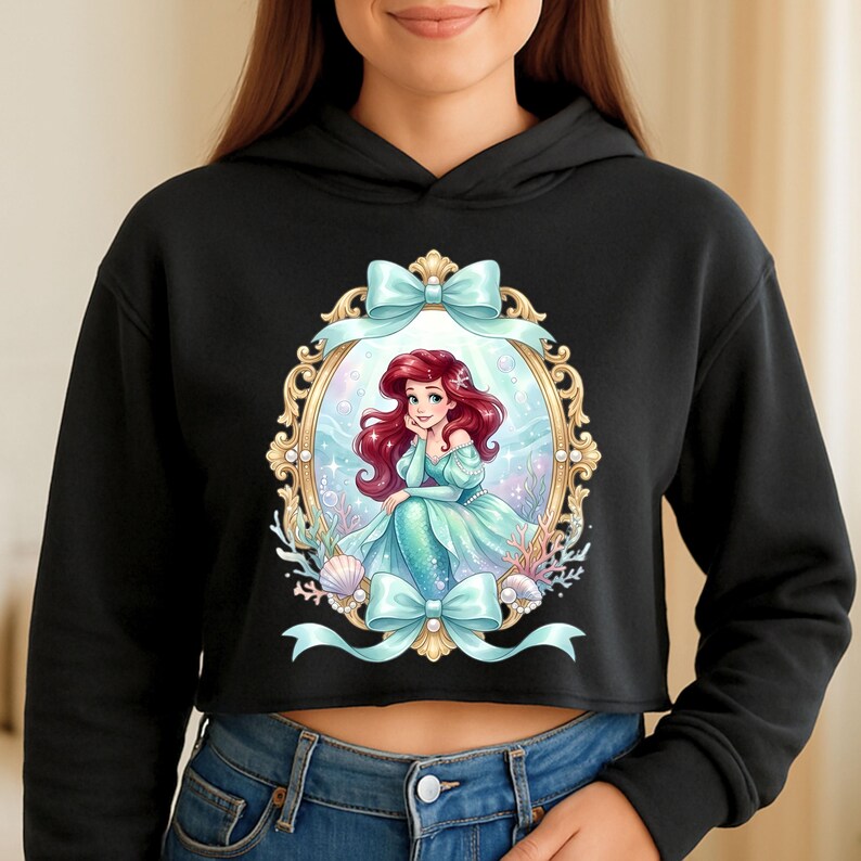Op de afbeelding: Zwarte cropped hoodie met een ingelijste illustratie van een zeemeermin met rood haar en een turquoise jurk. De lijst is goudkleurig met turquoise strikken. De zeemeermin is omringd door schelpen en koraal.