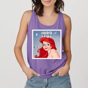 May include: A purple tank top featuring a graphic of Ariel from The Little Mermaid. The graphic includes the text "1989 Ariel's Version" with Ariel's image and seagulls against a blue sky background.
