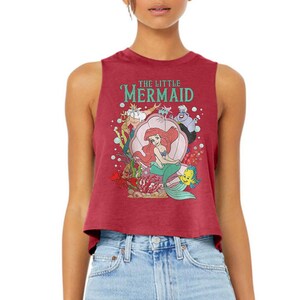 Puede incluir: Un top corto sin mangas de color granate con un estampado gr&aacute;fico de los personajes de La Sirenita, incluyendo a Ariel, el Rey Trit&oacute;n, &Uacute;rsula, Sebasti&aacute;n y Flounder. El texto THE LITTLE MERMAID est&aacute; impreso en color cerceta sobre los personajes.