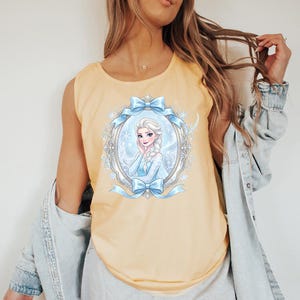 May include: A light yellow tank top featuring a framed illustration of a cartoon woman with a blue dress and a long braid. The frame is decorated with blue bows and snowflakes. The tank top is sleeveless.