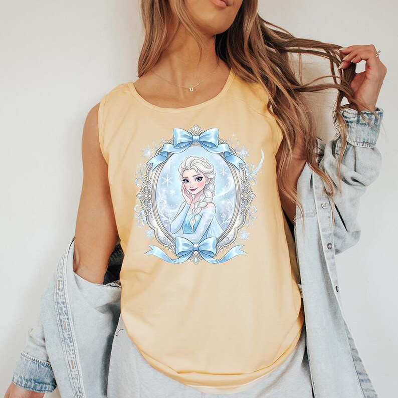 May include: A light yellow tank top featuring a framed illustration of a cartoon woman with a blue dress and a long braid. The frame is decorated with blue bows and snowflakes. The tank top is sleeveless.