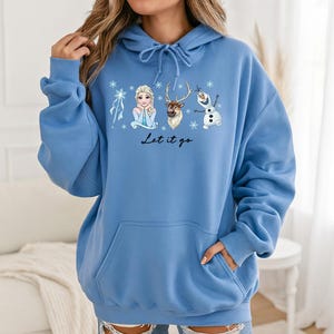 Op de afbeelding: Een lichtblauwe hoodie met een afbeelding van Elsa, Sven en Olaf uit Frozen, met sneeuwvlokken en de tekst "Let it go". De hoodie heeft een capuchon met trekkoord en een voorzak.