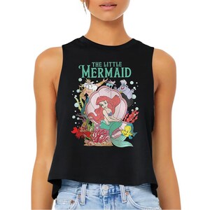 Puede incluir: Un top corto negro sin mangas con un estampado gr&aacute;fico de los personajes de La Sirenita, incluyendo a Ariel en una concha, el Rey Trit&oacute;n, &Uacute;rsula y Flounder. El texto THE LITTLE MERMAID est&aacute; impreso en color verde azulado sobre la escena.