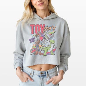 May include: A light grey cropped hoodie featuring a colourful graphic print of Toy Story characters Woody, Buzz Lightyear, and Rex. The design includes the text Toy Story, Since 1995, Woody, Buzz, and Rex, set against a background of geometric shapes.